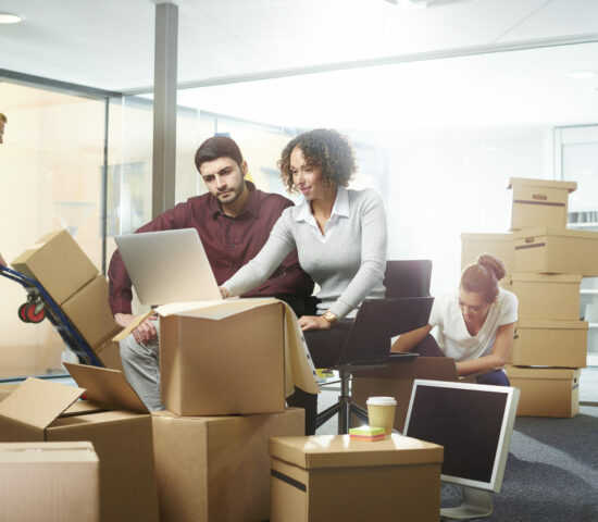 A business owner sits amongst his moving in boxes , waiting for the removal team to bring the desks . An interior designer is showing him the modifications needed as building contractor brings in various fixtures and fittings for the new office layout . They are looking at the layout plans and computer drawings . In the background her colleague is checking the files.