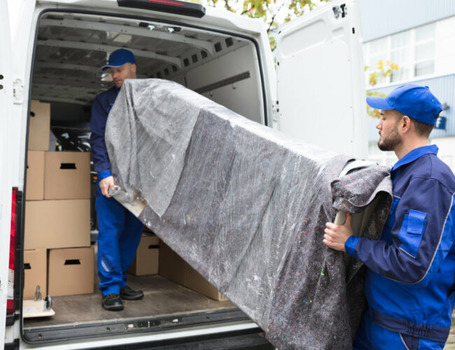 Two Young Delivery Men In Uniform Unloading Furniture From Vehicle